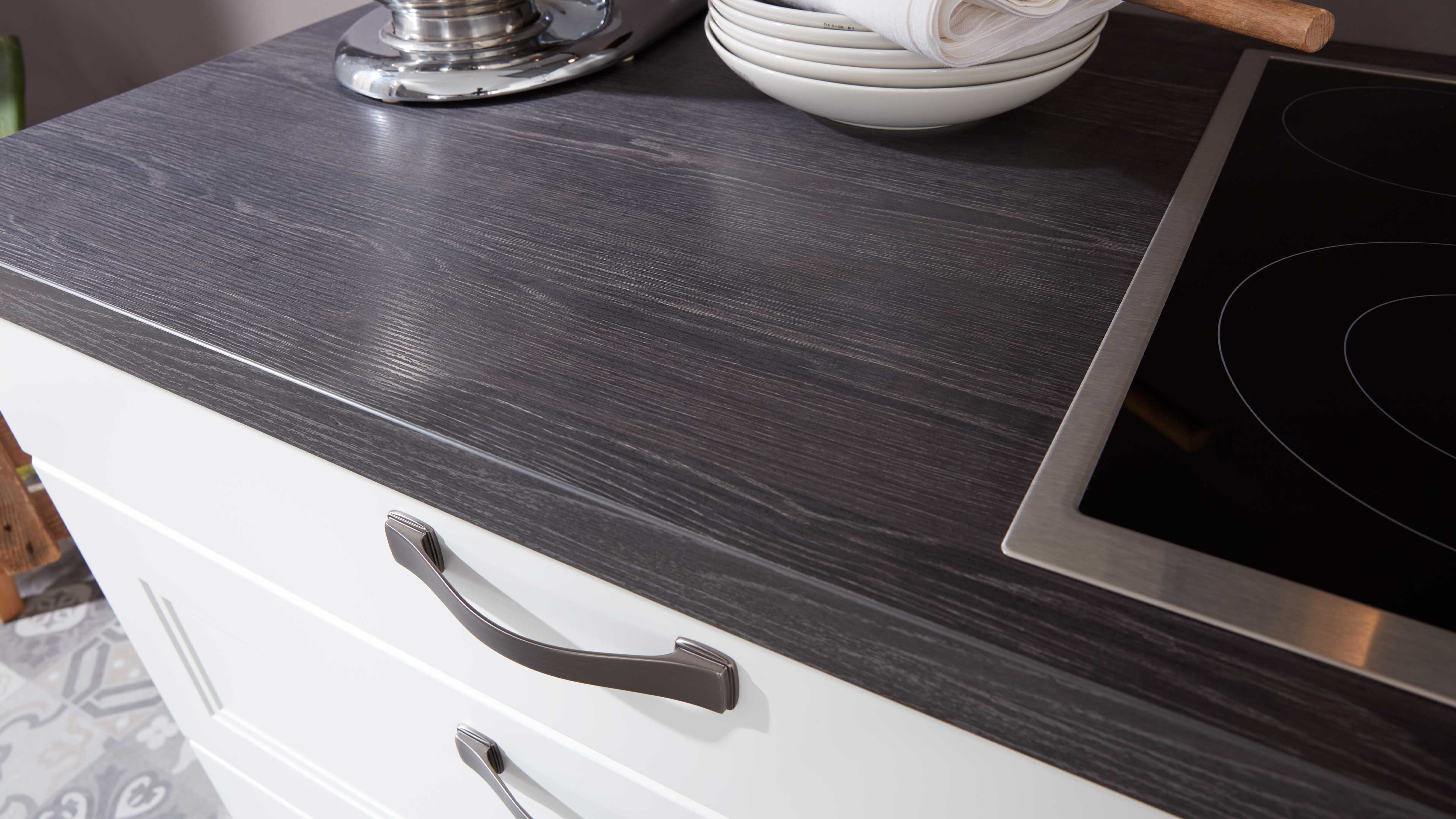 Küchenarbeitsplatte in dunkel gemasertem Holzdekor mit eingelassenem schwarzen Glaskochfeld und weißen Schubladen mit Metallgriffen.