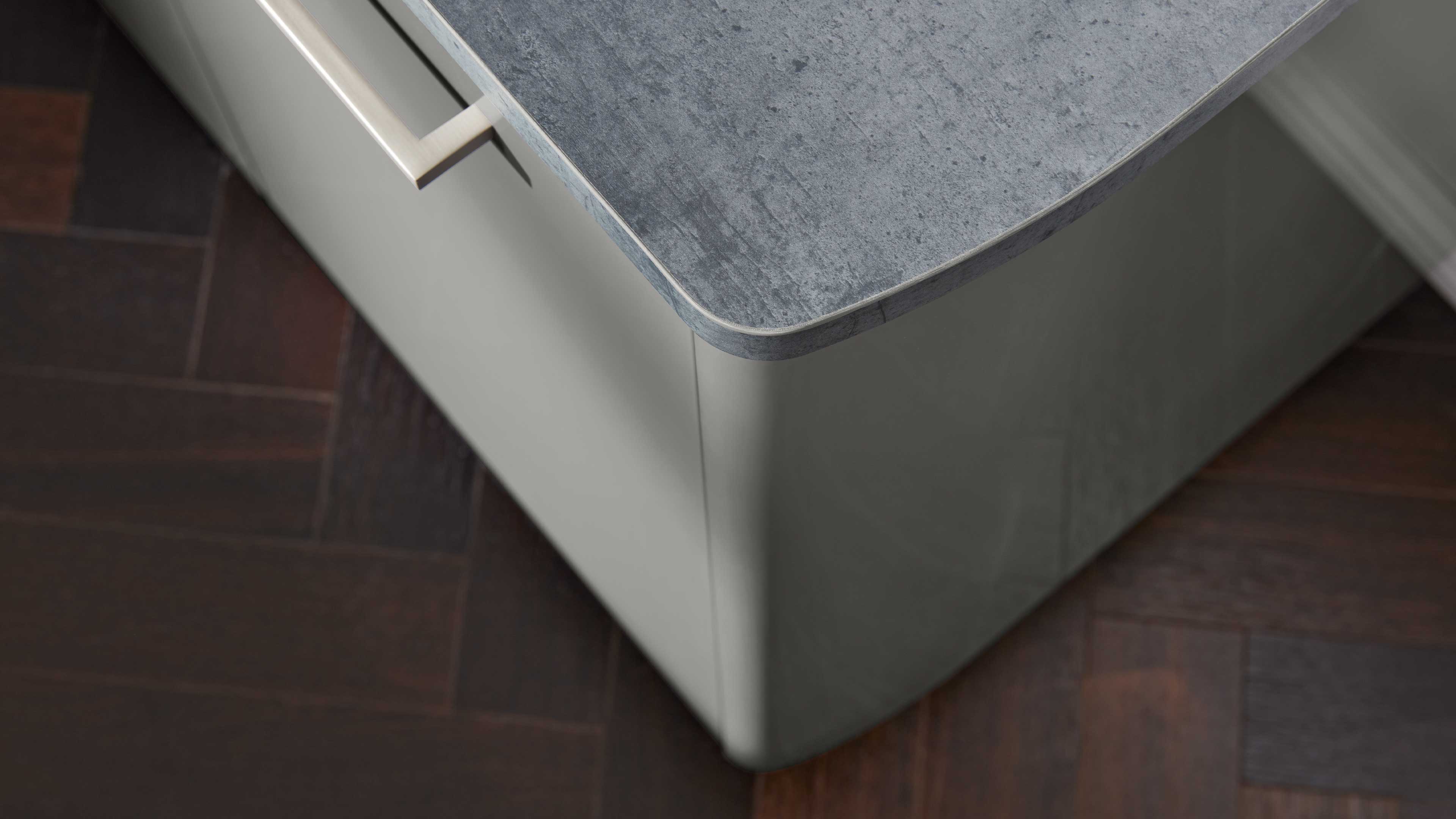 Eckdetail einer Arbeitsplatte in grauer Betonoptik mit abgerundeter Kante auf hellgrauem Schrank und Metallgriff.