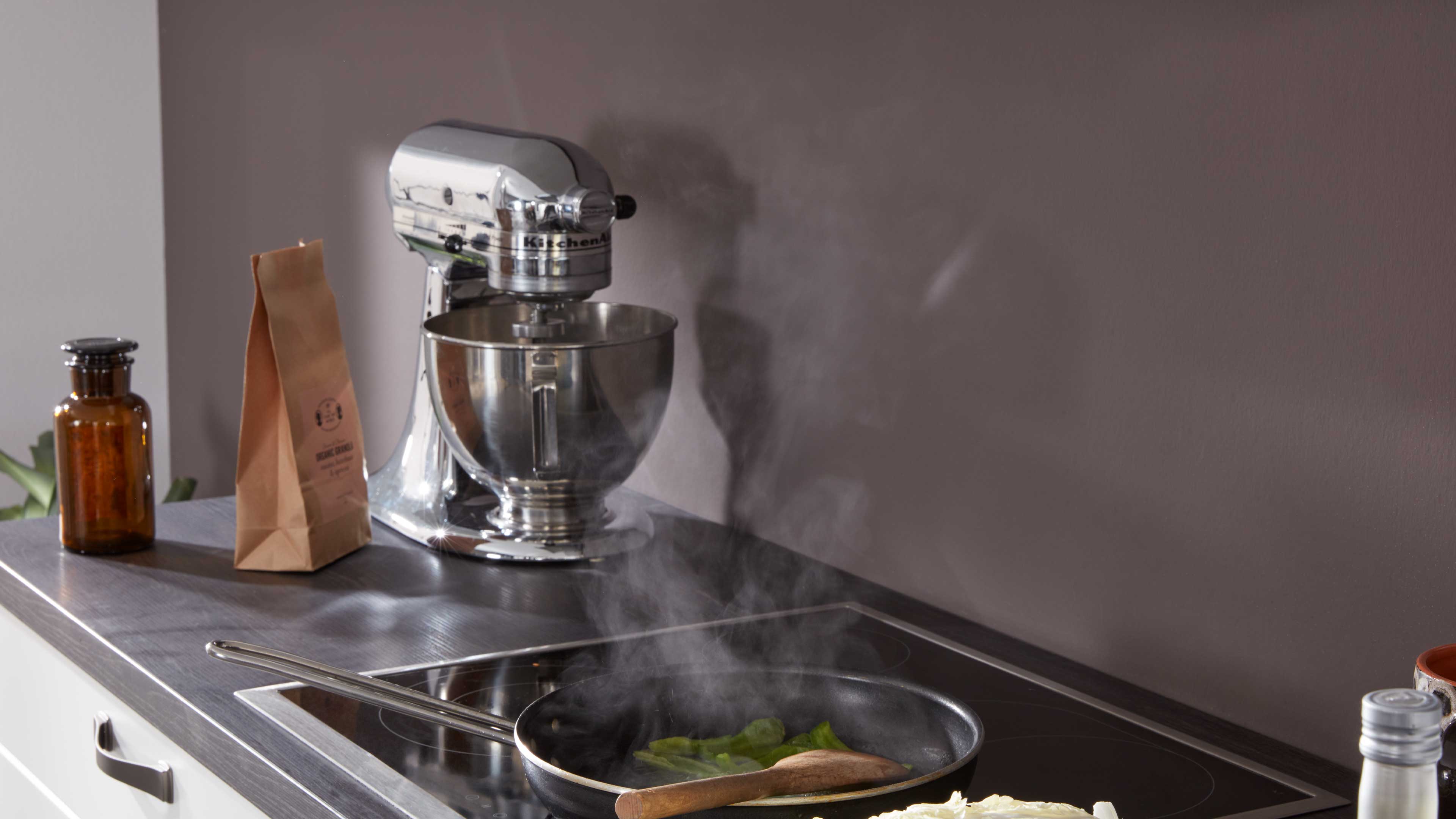 Chromfarbene Küchenmaschine aus Metall mit Rührschüssel auf dunkler Arbeitsplatte neben Induktionskochfeld und Pfanne.