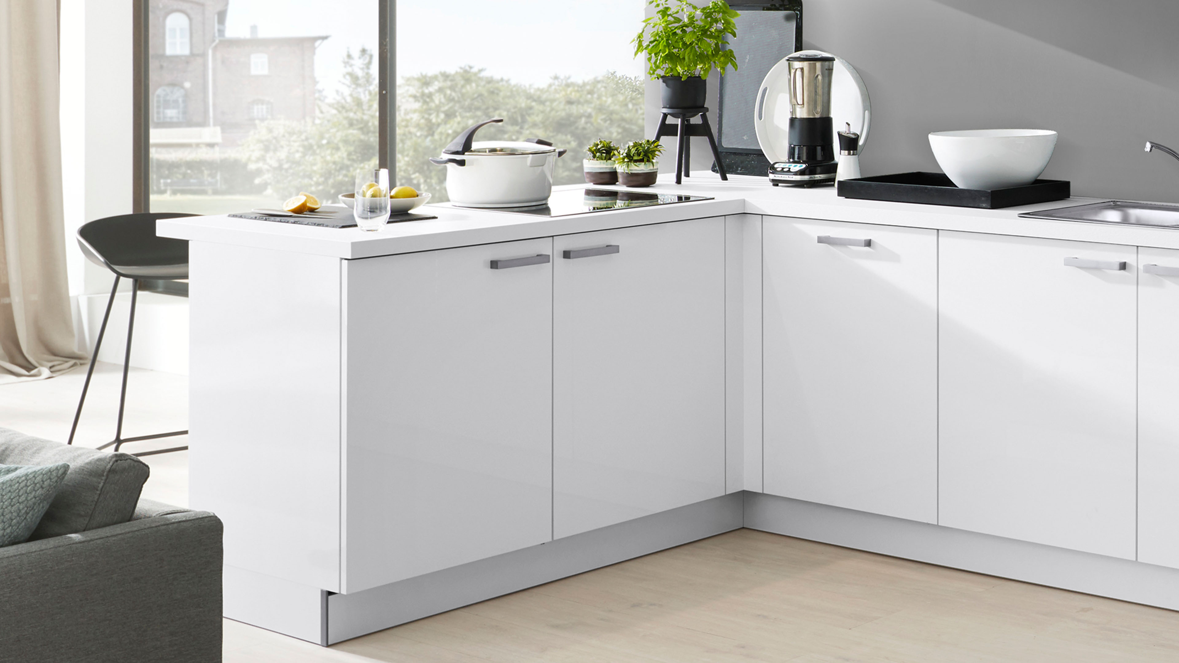 Weiße Eckküche mit hochglänzenden Fronten, rechteckigen silbernen Griffen, weißer Arbeitsplatte, eingelassenem Glaskochfeld und Edelstahlspüle.