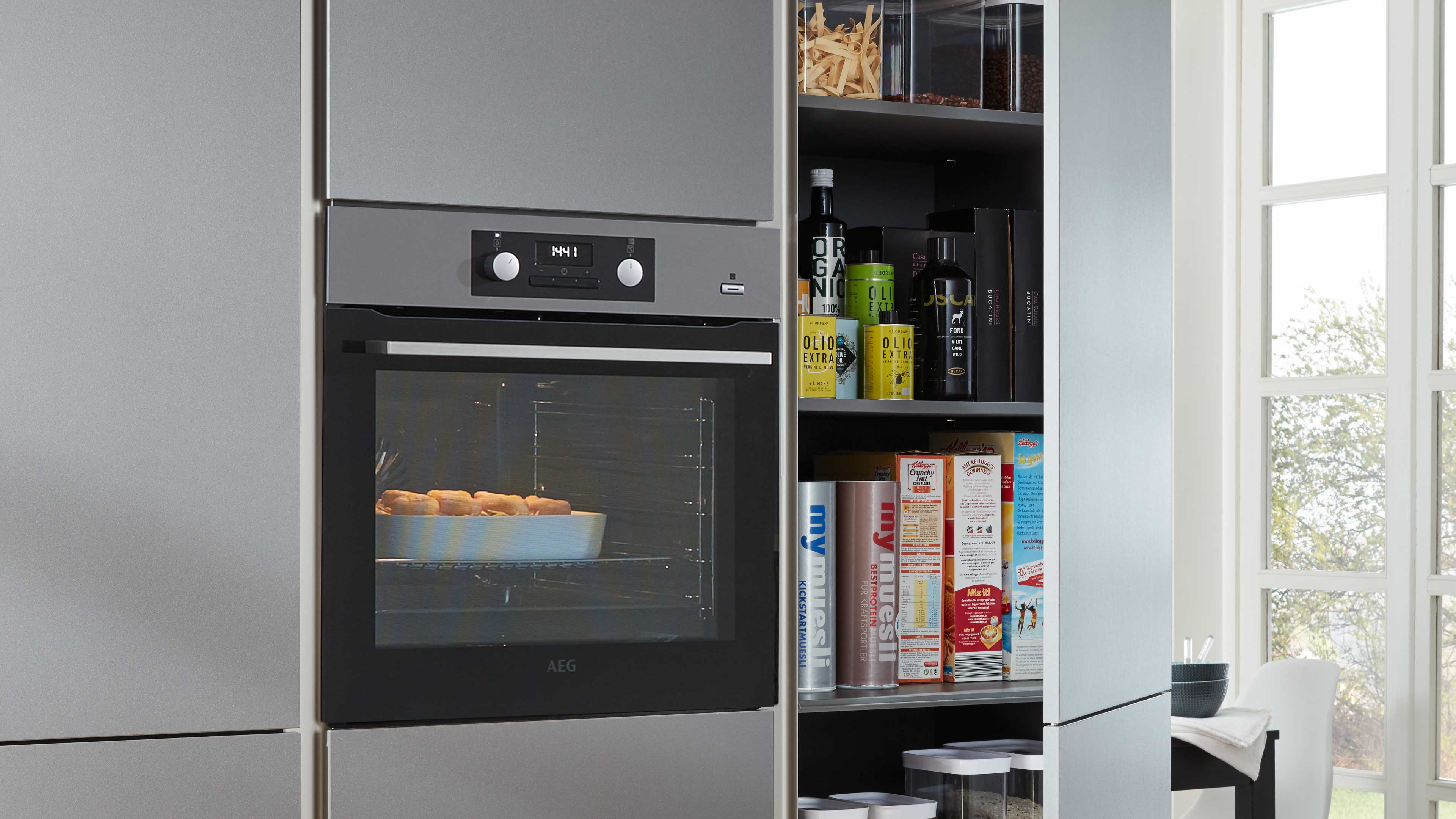Grauer Einbaubackofen mit sichtbarem Backblech und Gebäck, daneben offene Regalfächer mit Flaschen, Dosen und Müslipackungen.