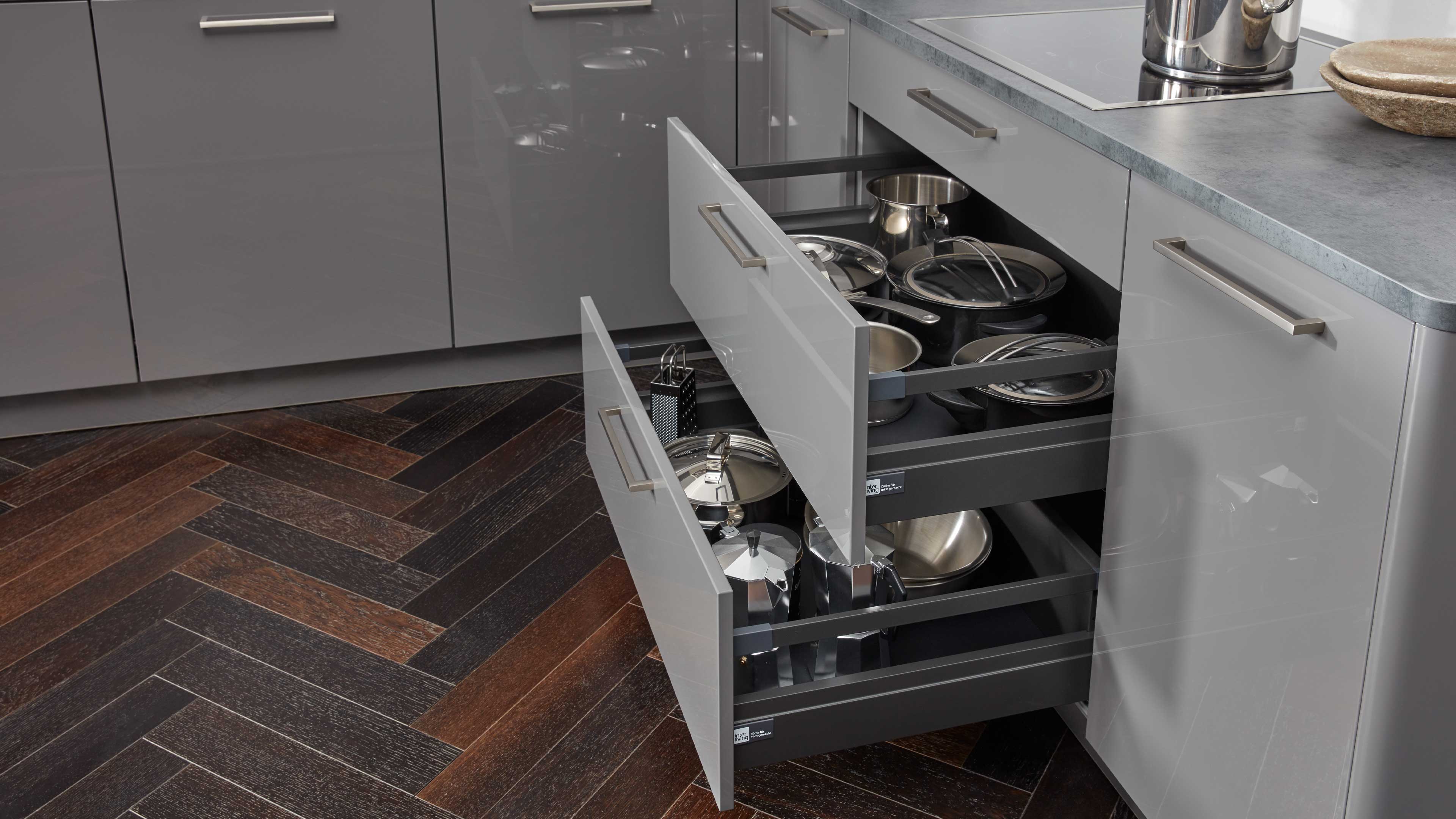 Zwei geöffnete graue Küchenschubladen mit metallenen Griffen, schwarzem Innenraum und eingeordneten Edelstahl-Töpfen, Pfannen und Deckeln.