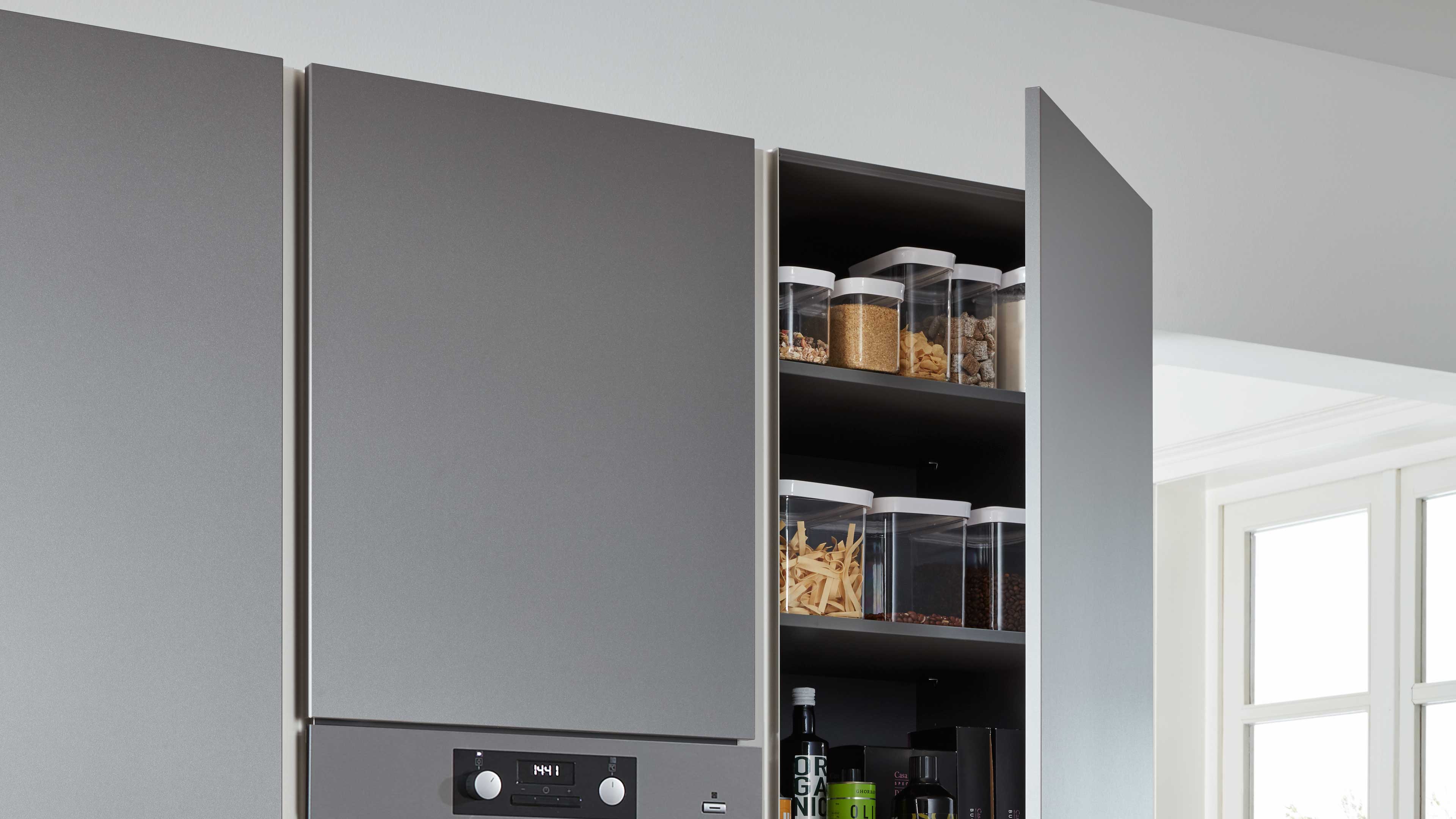 Hoher Wandschrank mit matten grauen Fronten, eine Tür offen, innen dunkle Regalböden mit transparenten Vorratsdosen und Flaschen.