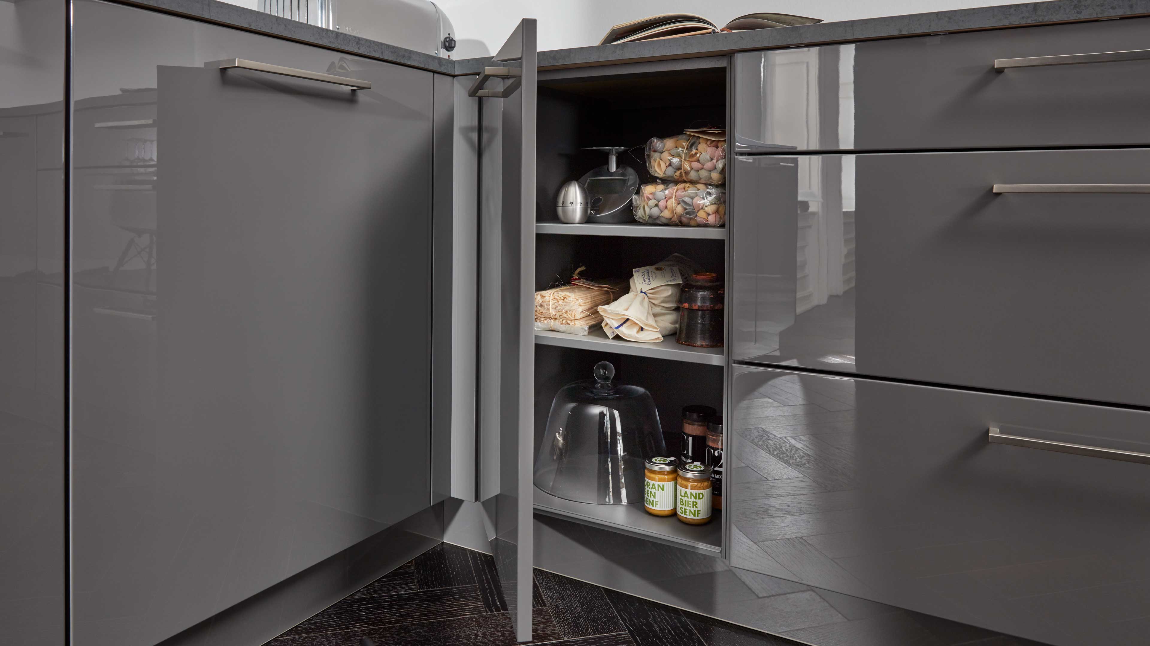 Eckunterschrank mit geöffneter Tür, graue Hochglanzfronten und Metallgriffe, drei Regalböden mit Glashaube, Gläsern, Dosen und Packungen.
