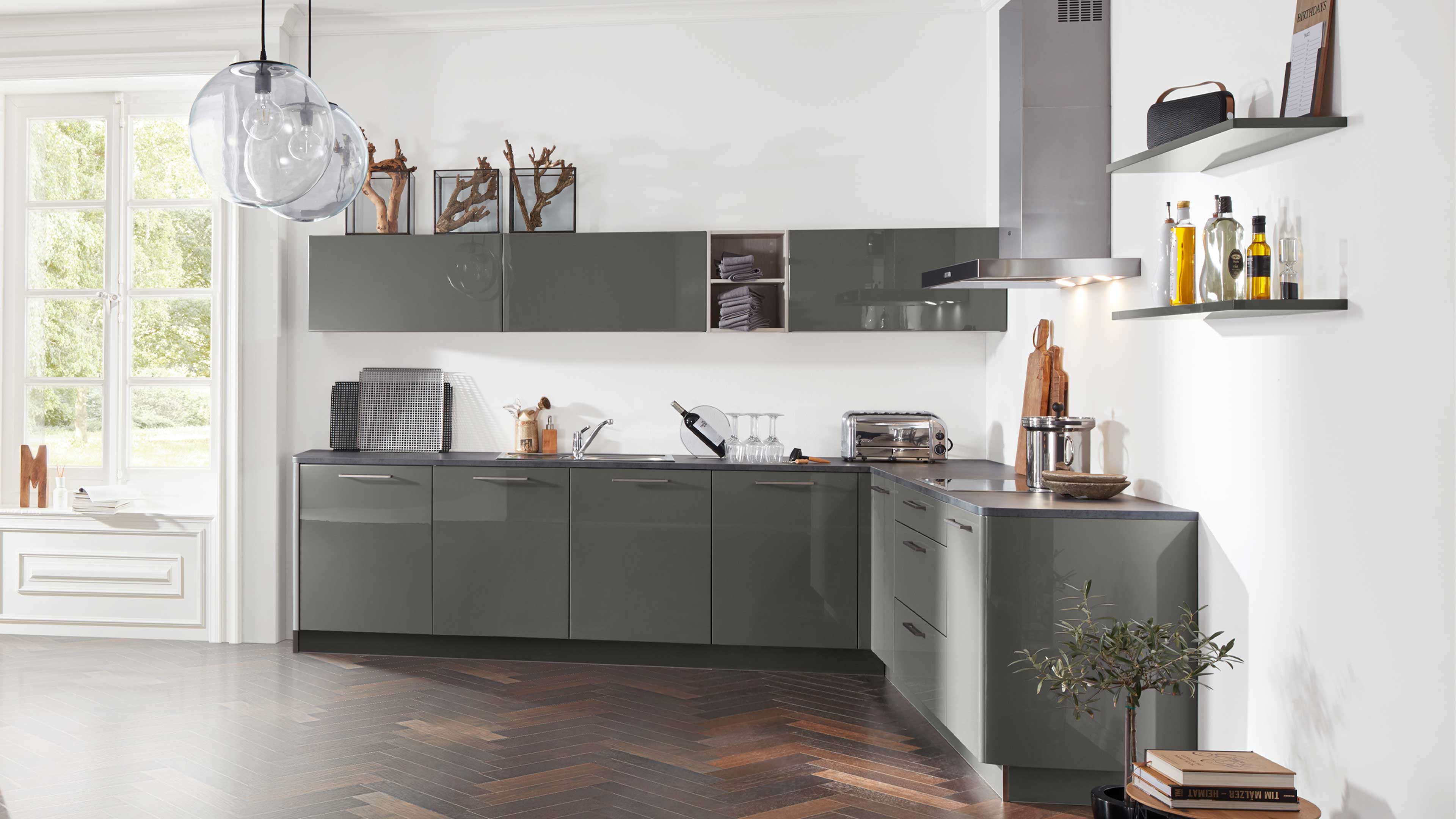 L-förmige Einbauküche mit hochglänzenden graugrünen Fronten, dunkler Arbeitsplatte, Edelstahlhaube und Glas-Leuchte.