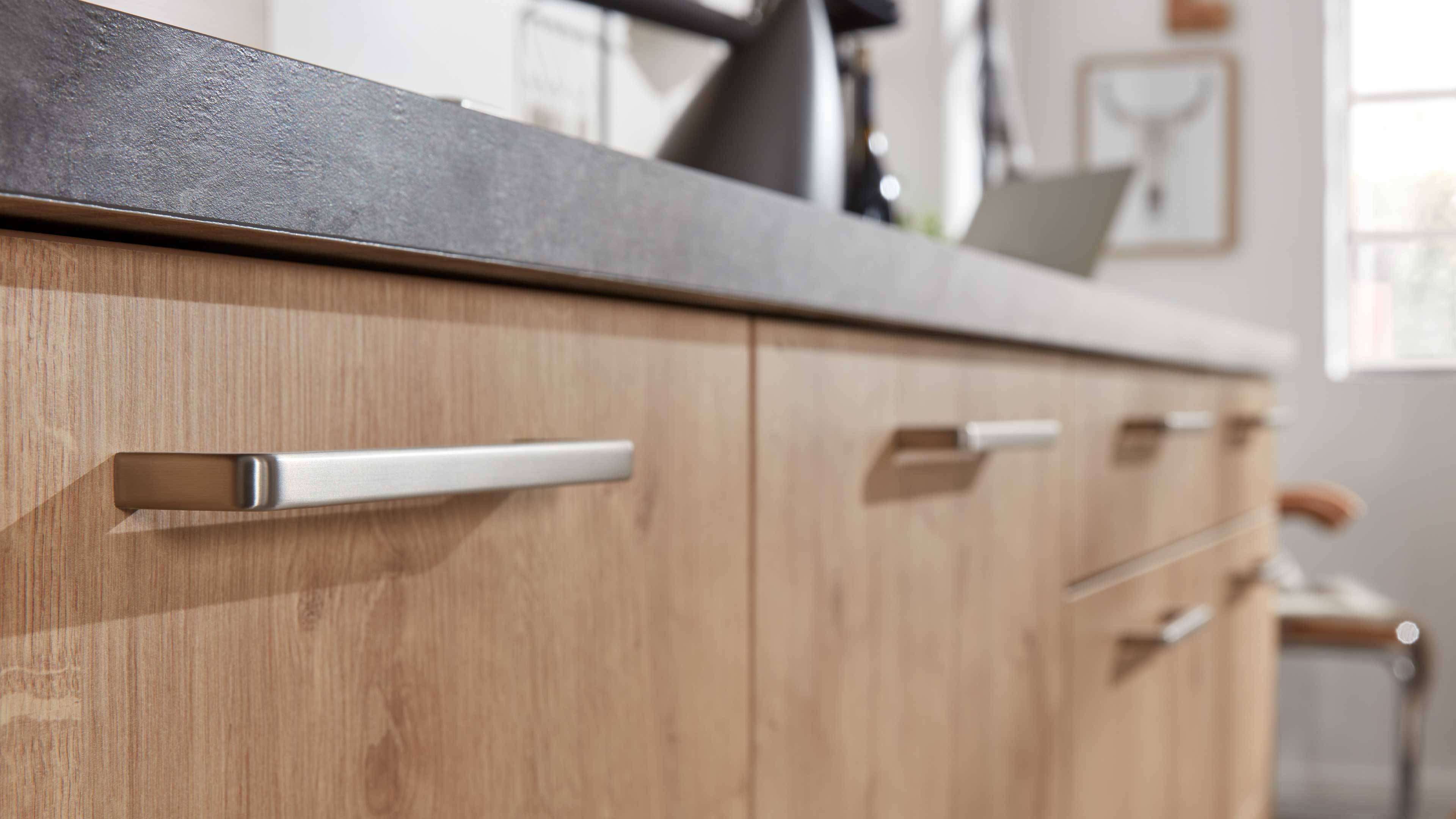 Küchenfront mit hellholzoptik, schlichten länglichen Metallgriffen und einer dunklen, steinähnlichen Arbeitsplatte.