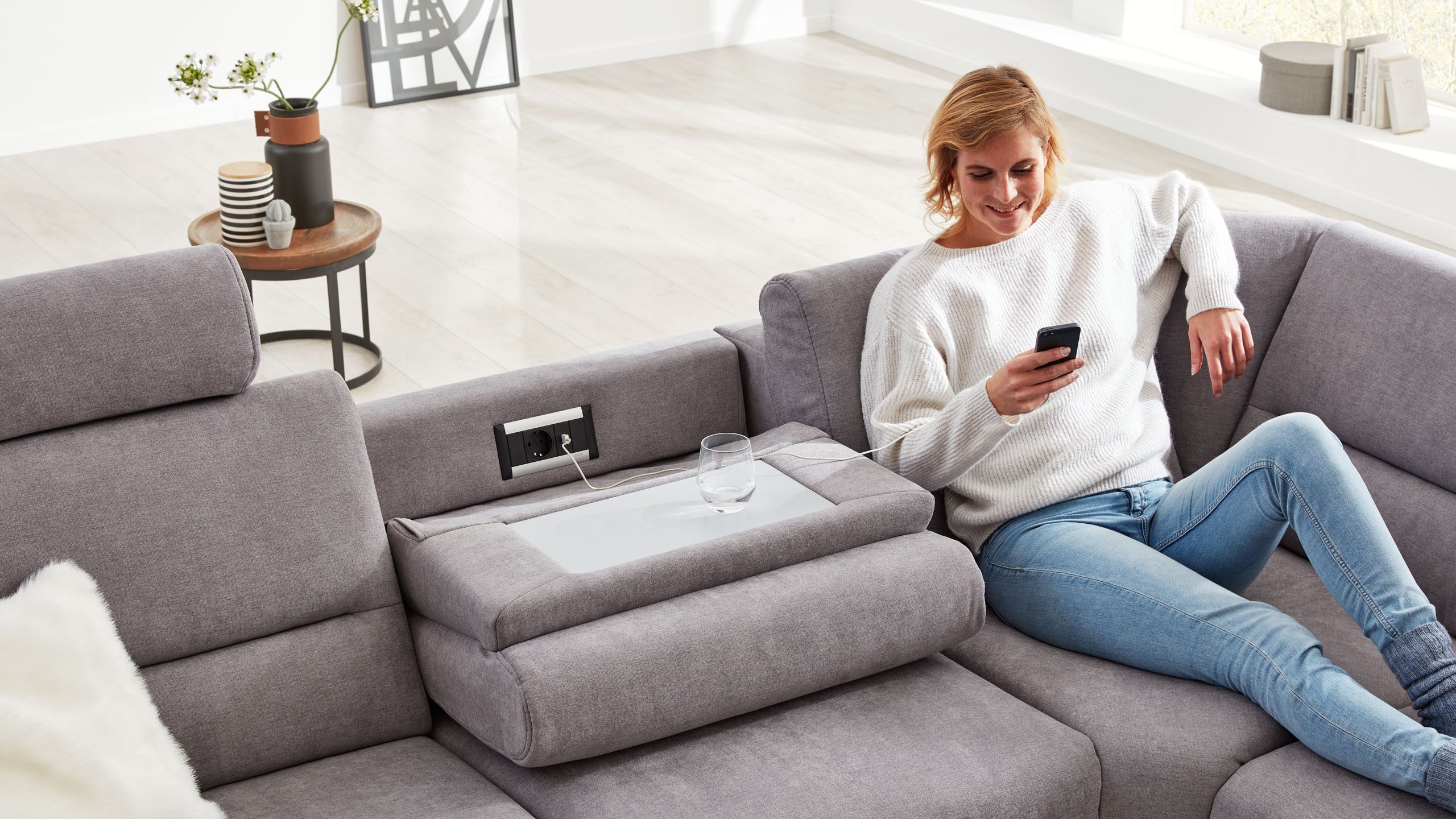 Frau liegt auf dem Sofa der Interliving Serie 4305 und lädt ihr Smartphone an einer integrierten Steckdose
