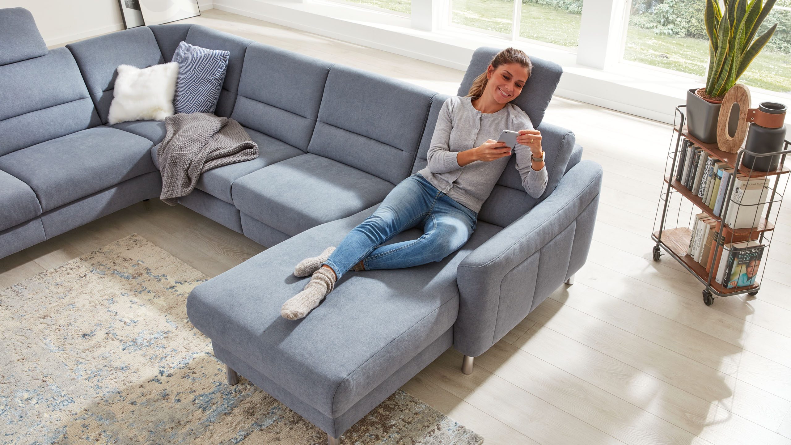 Frau surft in ihrem Smartphone und liegt auf dem Interliving Sofa Serie 4305